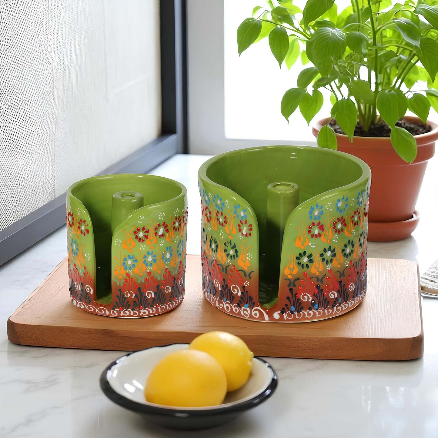 Two colorful ceramic paper towel holders with floral designs on a wooden board, with a bowl of lemons and a potted plant in the background.