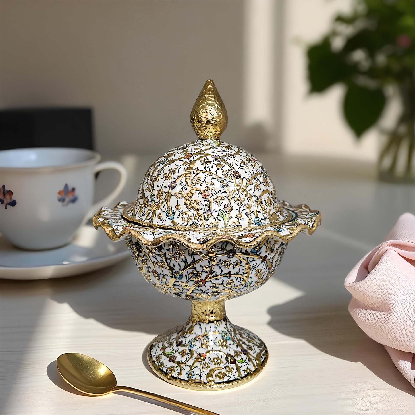 Decorative gold and silver covered sugar bowl on a table with a cup and spoon in the background