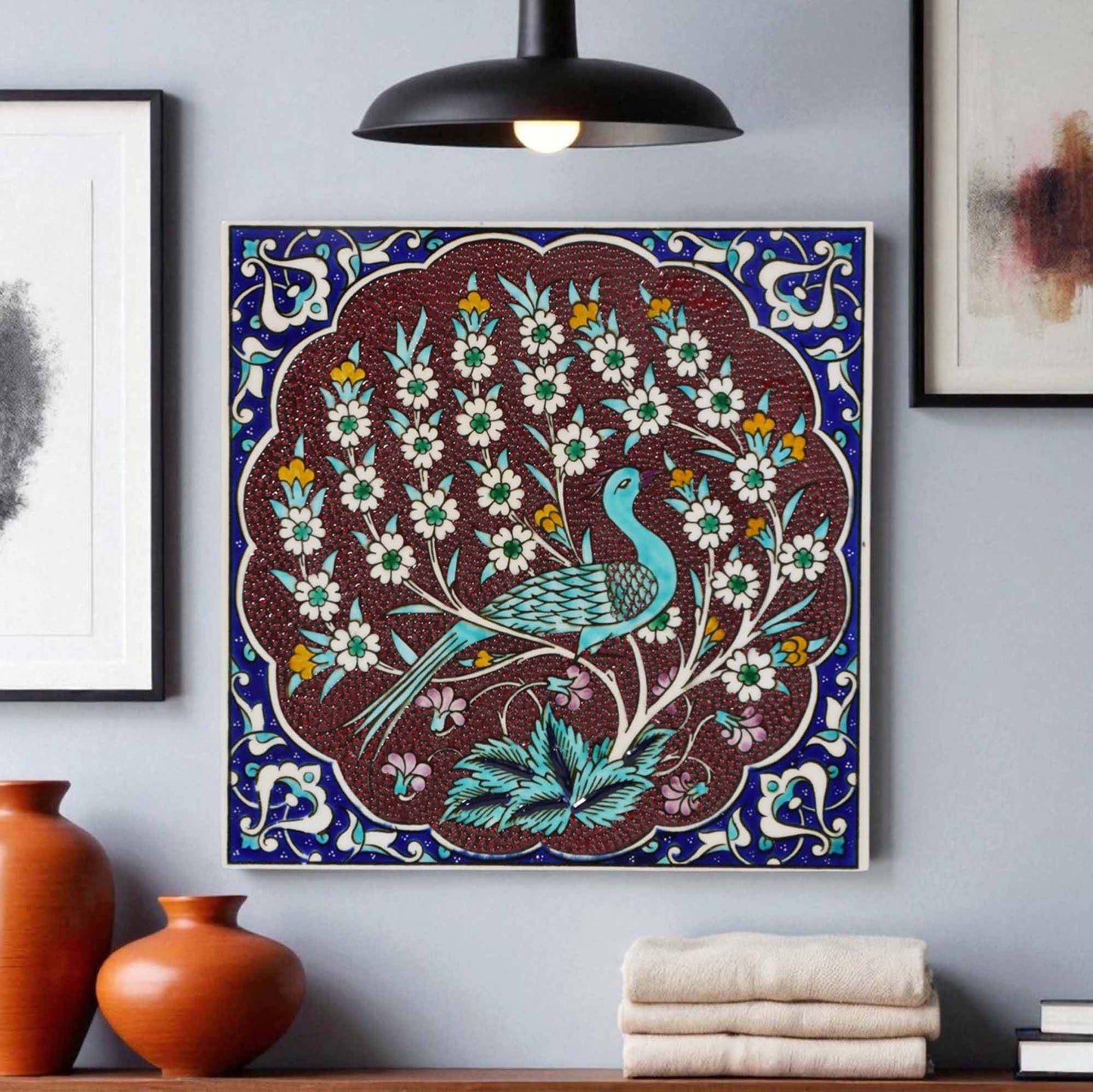 Decorative ceramic tile wall art with a peacock design on a blue background, hanging on a gray wall with vases and books below.