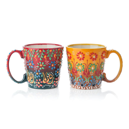 Two handcrafted ceramic coffee mugs, one in red and one in honey yellow, both with colorful floral patterns.
