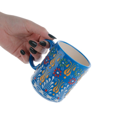 A hand holding a colorful handcrafted ceramic coffee mug with floral patterns.