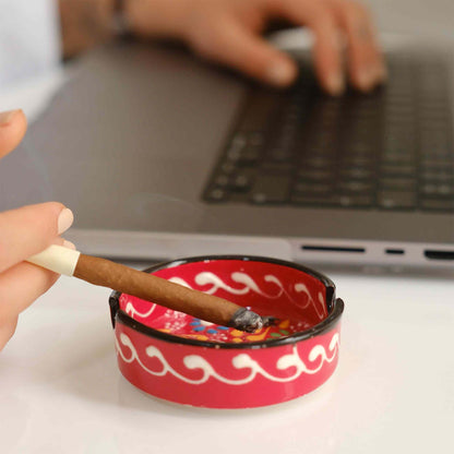 Person holding a lit cigar over a red ceramic ashtray with a laptop in the background