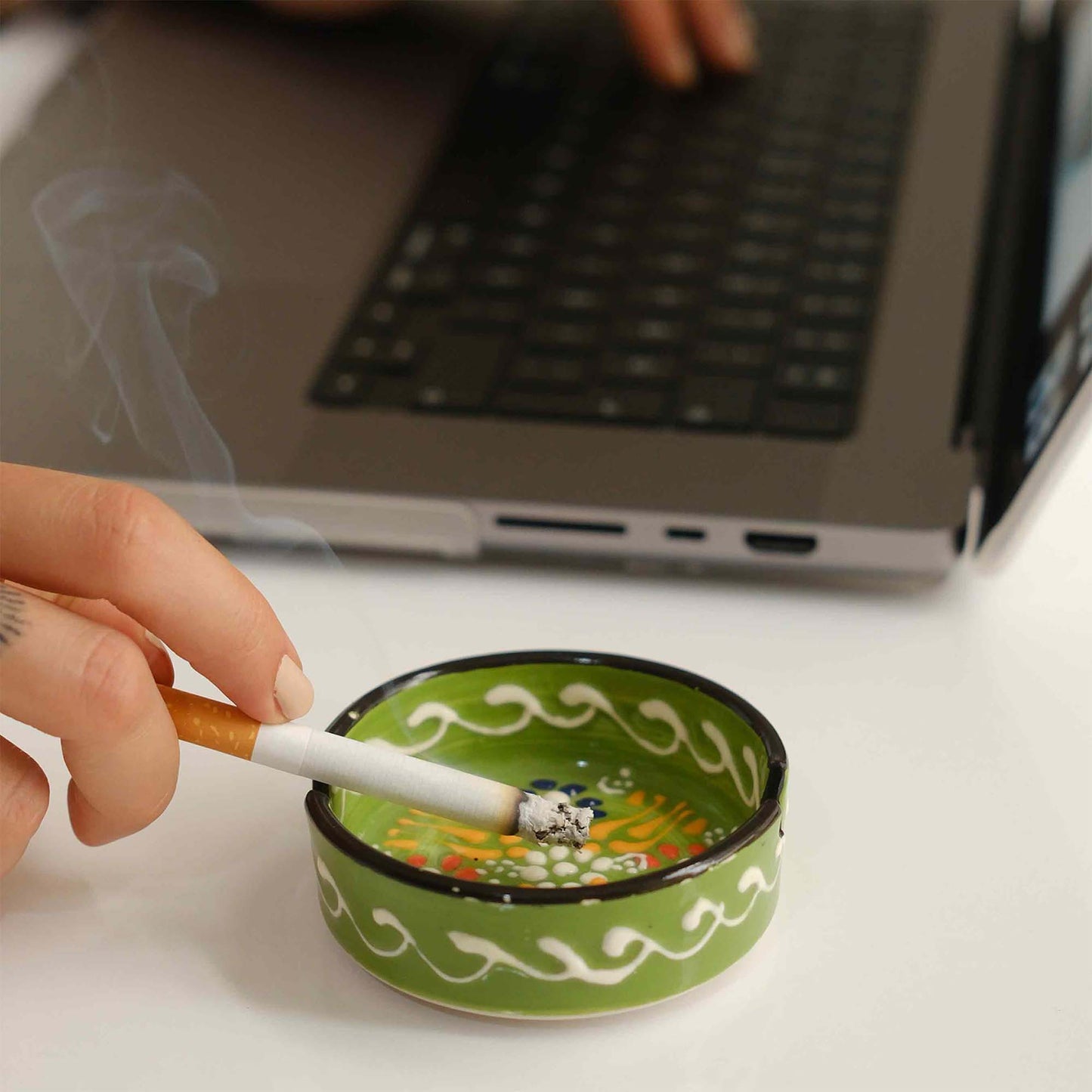 Person smoking a cigarette over a green ceramic ashtray with a laptop in the background