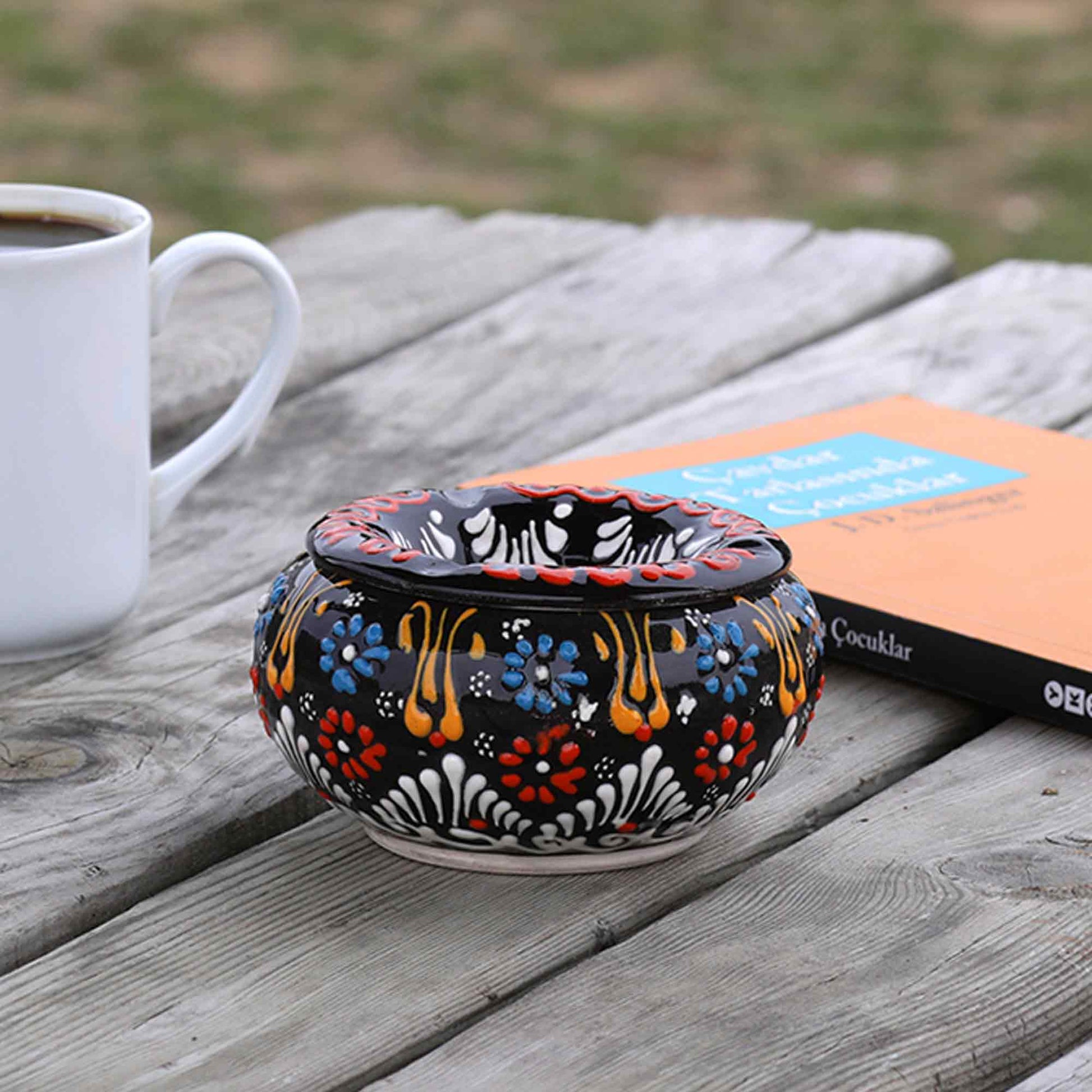 Decorative ceramic ashtray with floral patterns on a wooden surface next to a white mug.