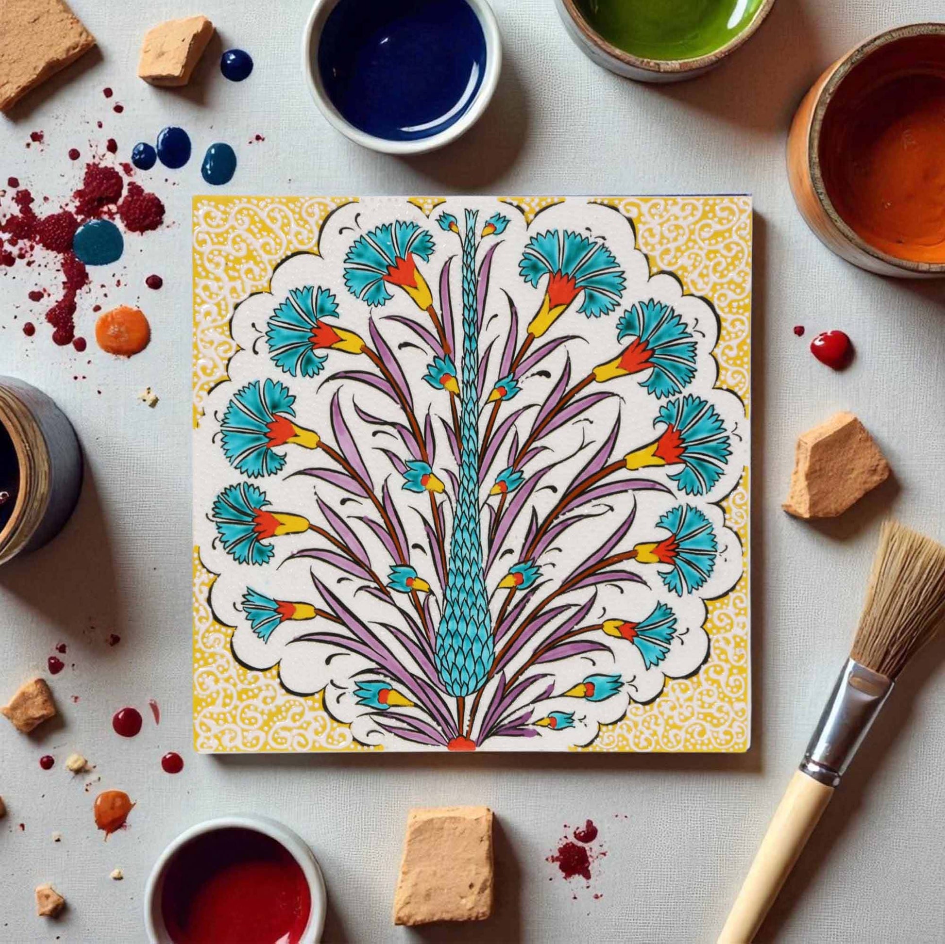 Decorative ceramic tile with floral design surrounded by paint containers and a brush on a light surface.