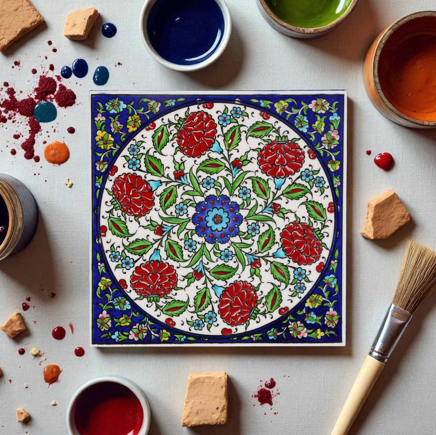 Decorative ceramic tile with floral pattern surrounded by paint containers and a brush on a light surface.