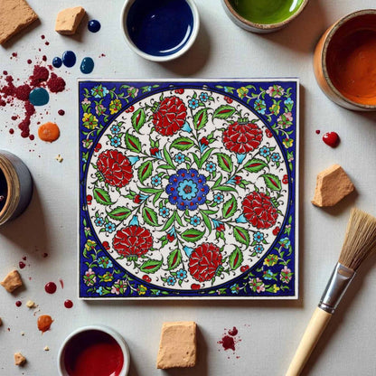 Decorative ceramic tile with floral pattern surrounded by paint containers and a brush on a light surface.