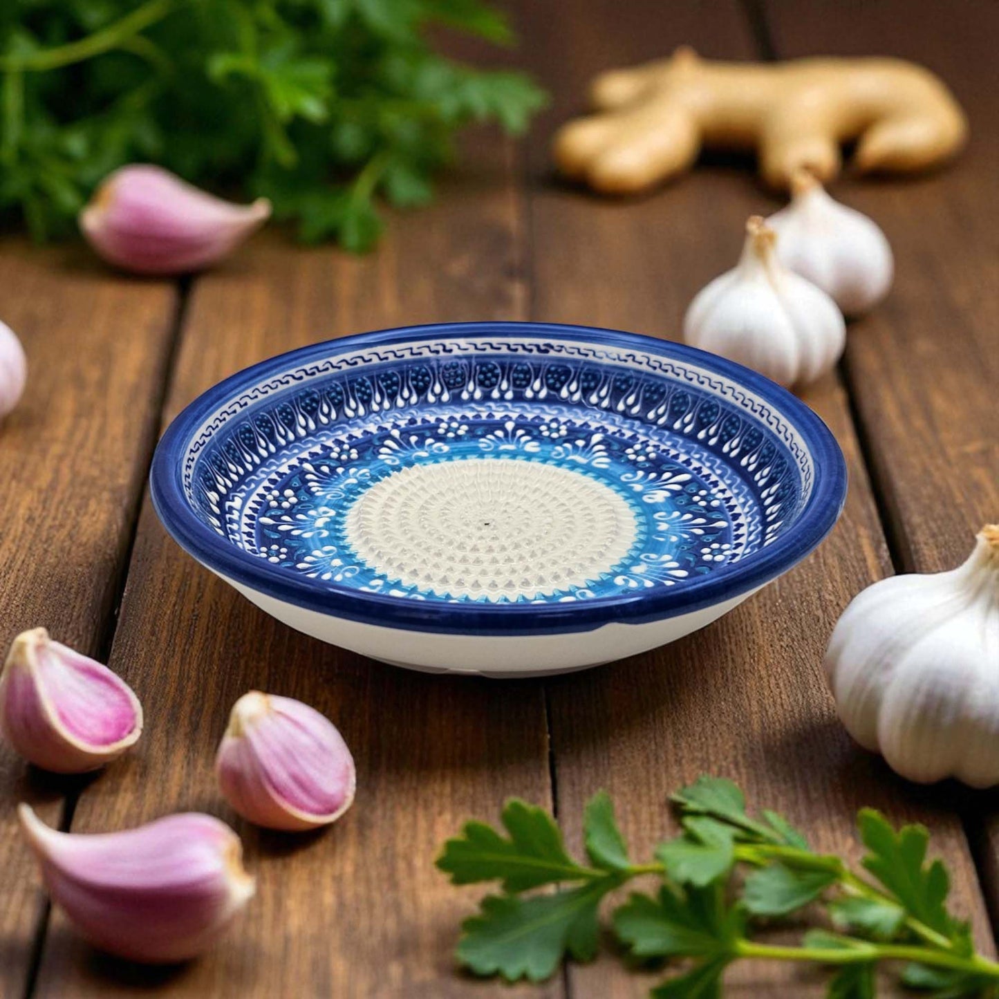 Blue and white decorative garlic grater bowl on a wooden surface with garlic and herbs.