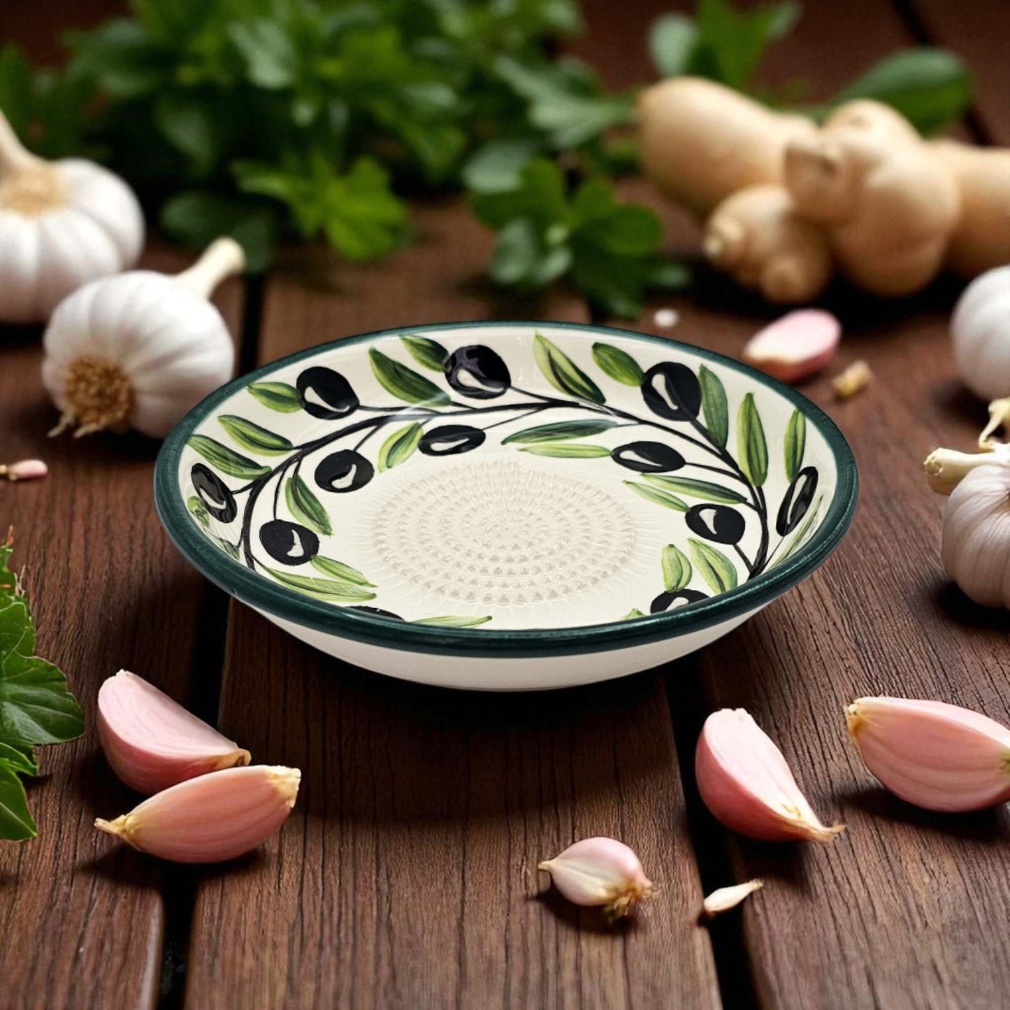 Decorative ceramic garlic grater bowl with olive design on a wooden surface with garlic and herbs.