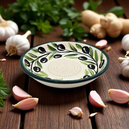 Decorative ceramic garlic grater bowl with olive design on a wooden surface with garlic and herbs.