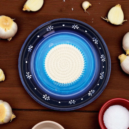 Blue ceramic garlic grater bowl with white center on a wooden surface surrounded by garlic bulbs.