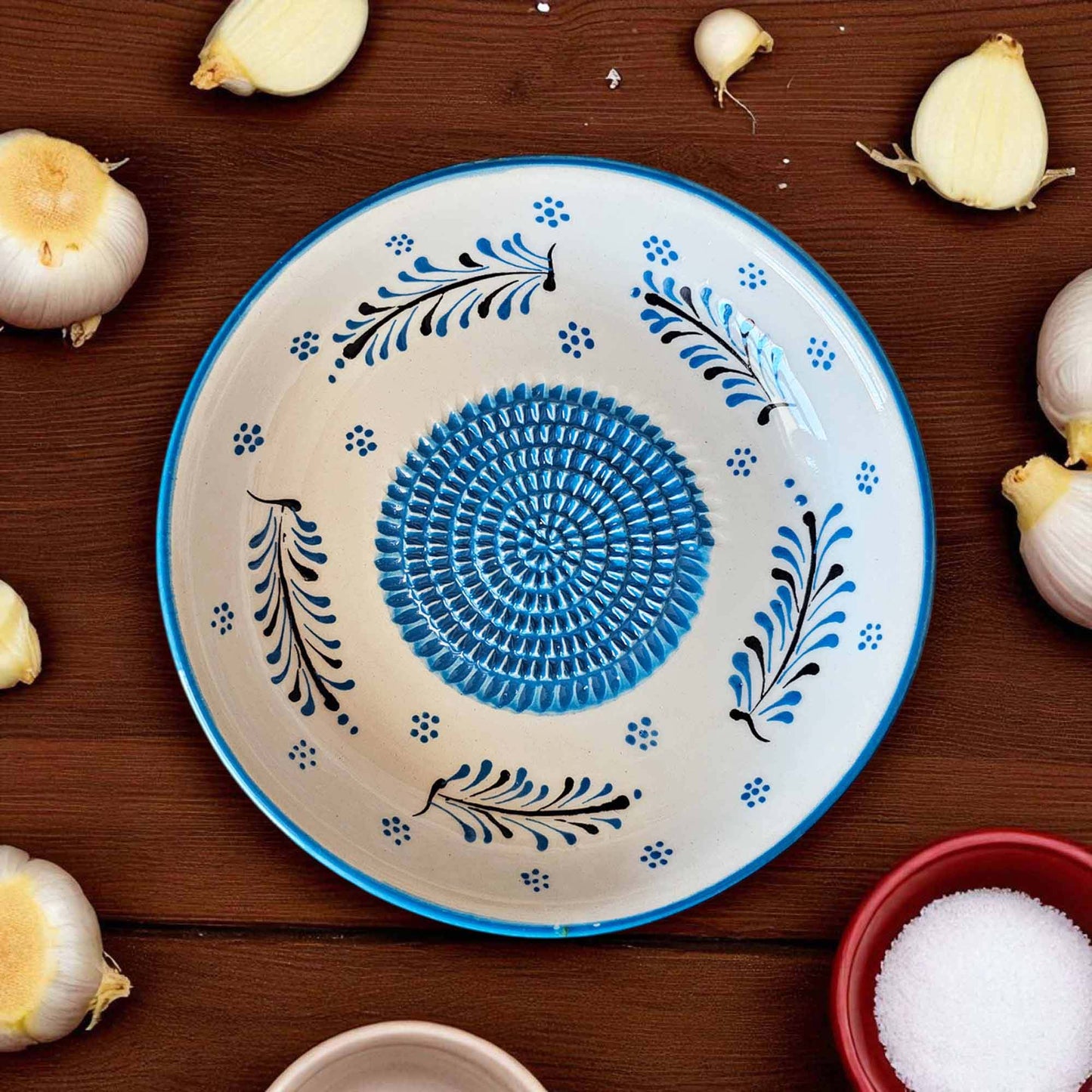 Decorative ceramic garlic grater bowl with blue and black patterns on a wooden surface with garlic bulbs.