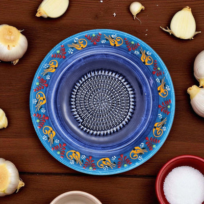 Decorative blue garlic grater bowl with colorful patterns on a wooden surface with garlic bulbs and a bowl of salt.