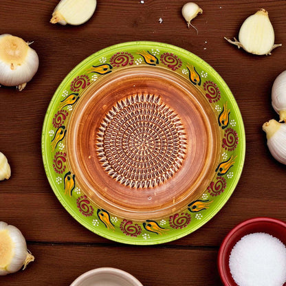 Decorative ceramic garlic grater bowl with green rim and brown center on a wooden surface with garlic bulbs.