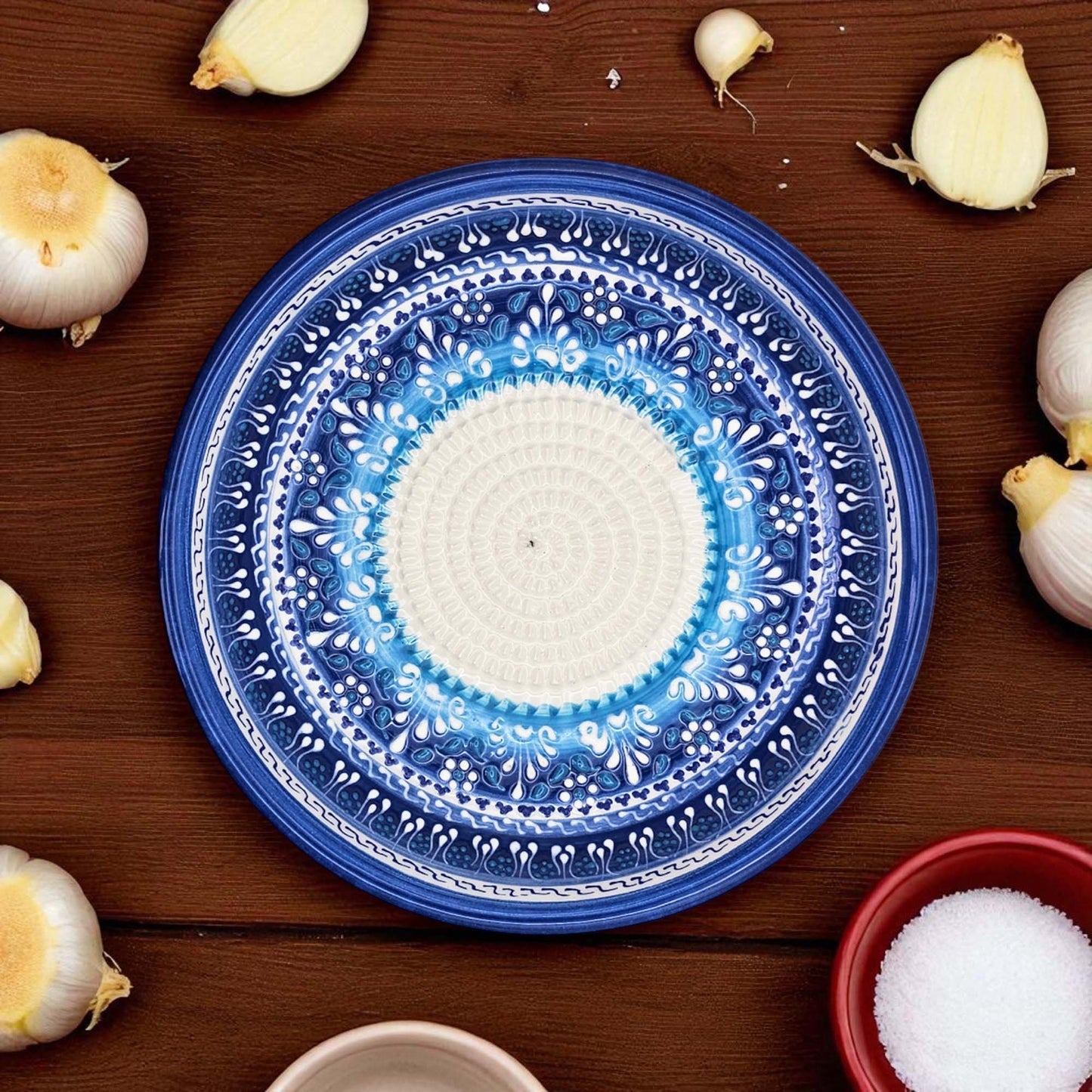 Blue decorative garlic grater bowl with white center on a wooden surface surrounded by garlic bulbs and a bowl of salt.