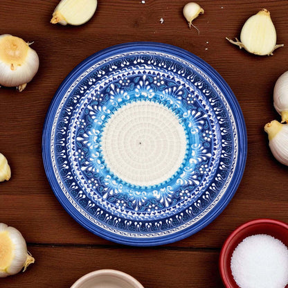 Blue decorative garlic grater bowl with white center on a wooden surface surrounded by garlic bulbs and a bowl of salt.