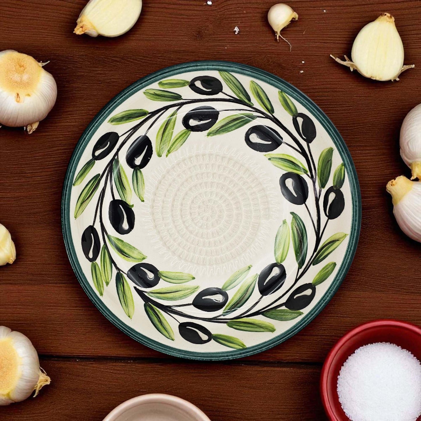 Decorative garlic grater bowl with olive design on a wooden surface with garlic bulbs and a bowl of salt.