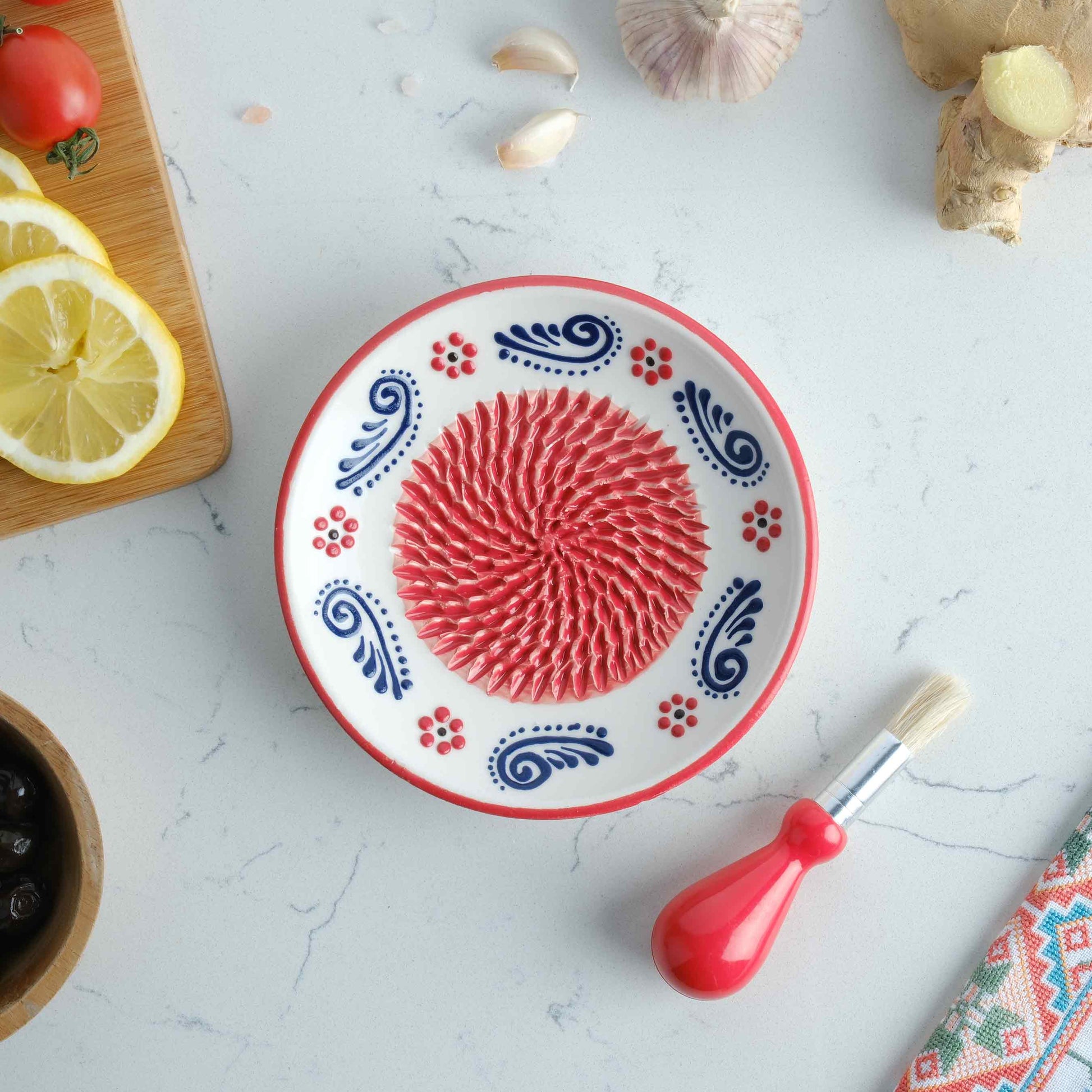 Decorative ceramic garlic grater plate with red central design on a white surface with kitchen items.