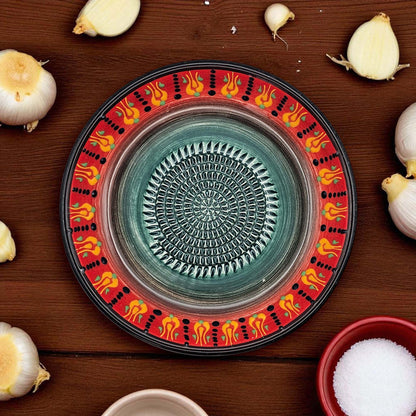 Decorative garlic grater bowl with a colorful rim on a wooden surface with garlic bulbs and a bowl of salt.