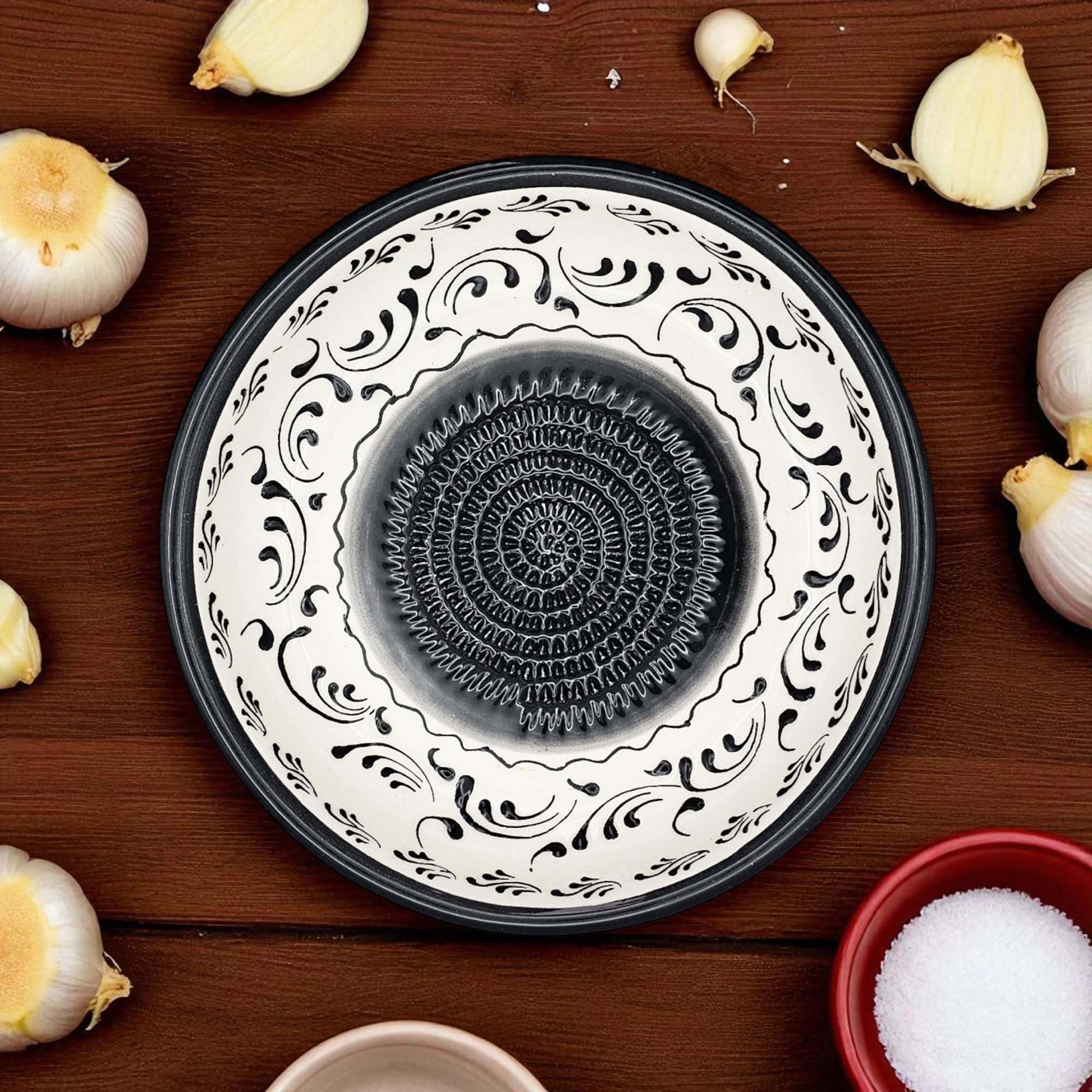 Decorative garlic grater bowl with black and white patterns on a wooden surface with garlic bulbs.