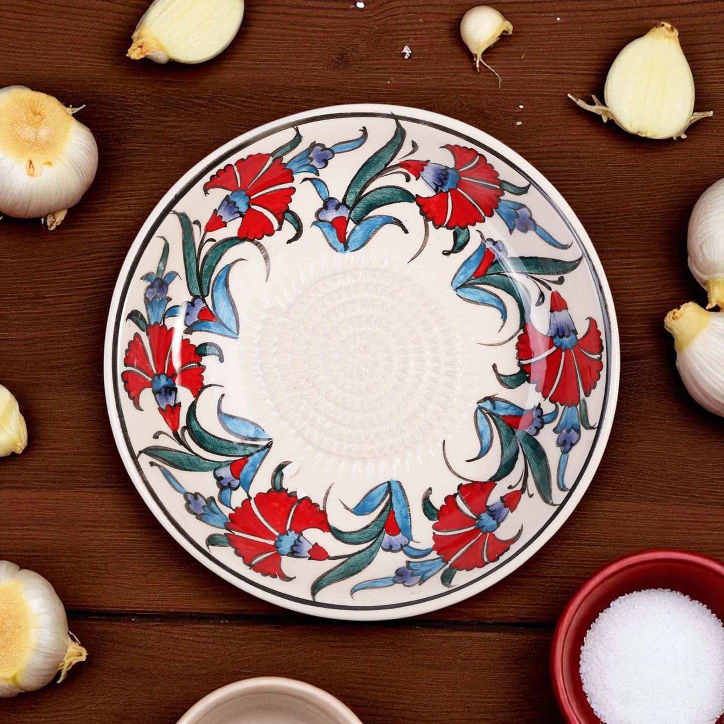 Decorative ceramic garlic grater bowl with floral design on a wooden surface with garlic bulbs.