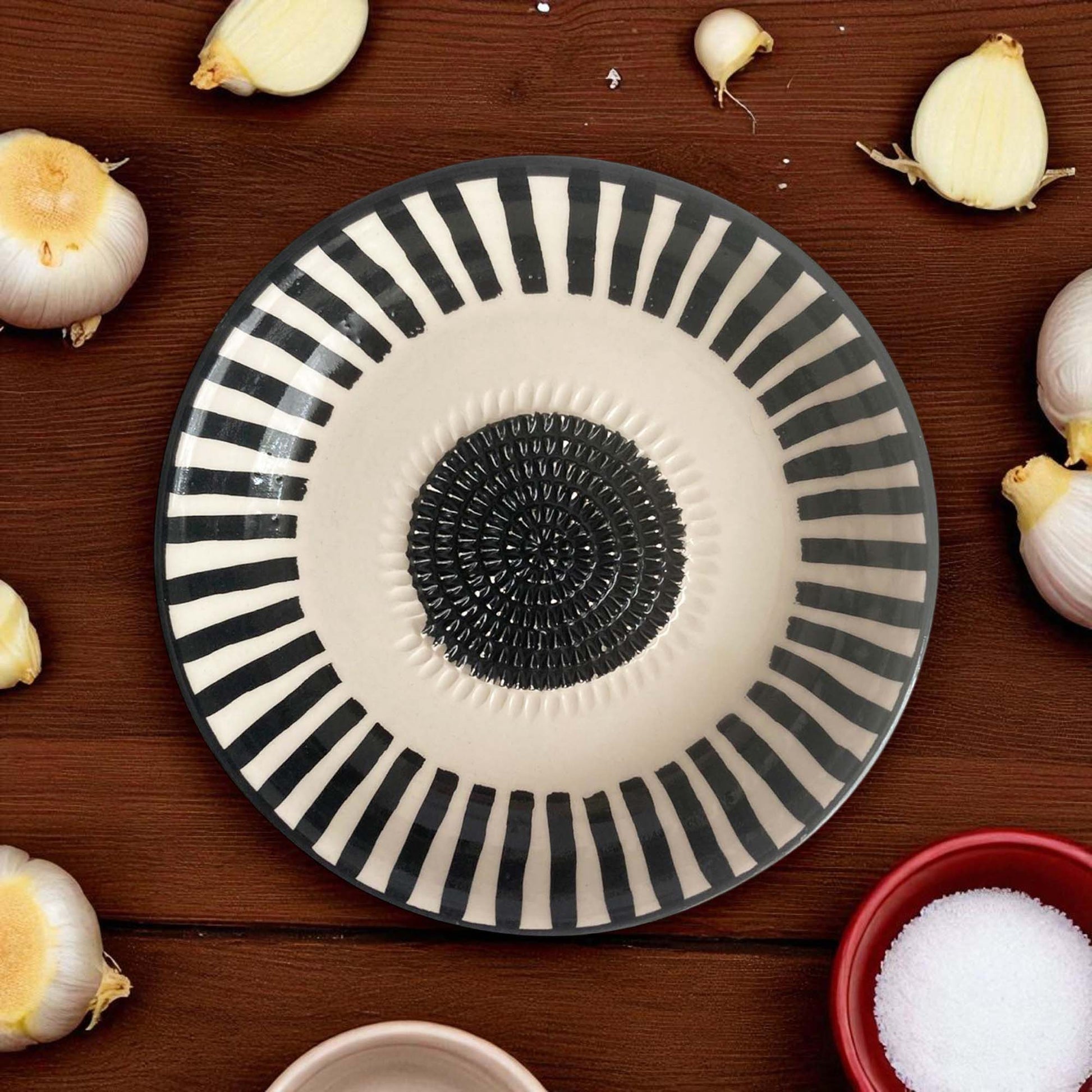 Decorative garlic grater bowl with black and white stripes on a wooden surface with garlic bulbs.