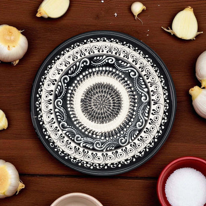 Decorative black and white garlic grater bowl on a wooden surface with garlic bulbs and a bowl of salt.