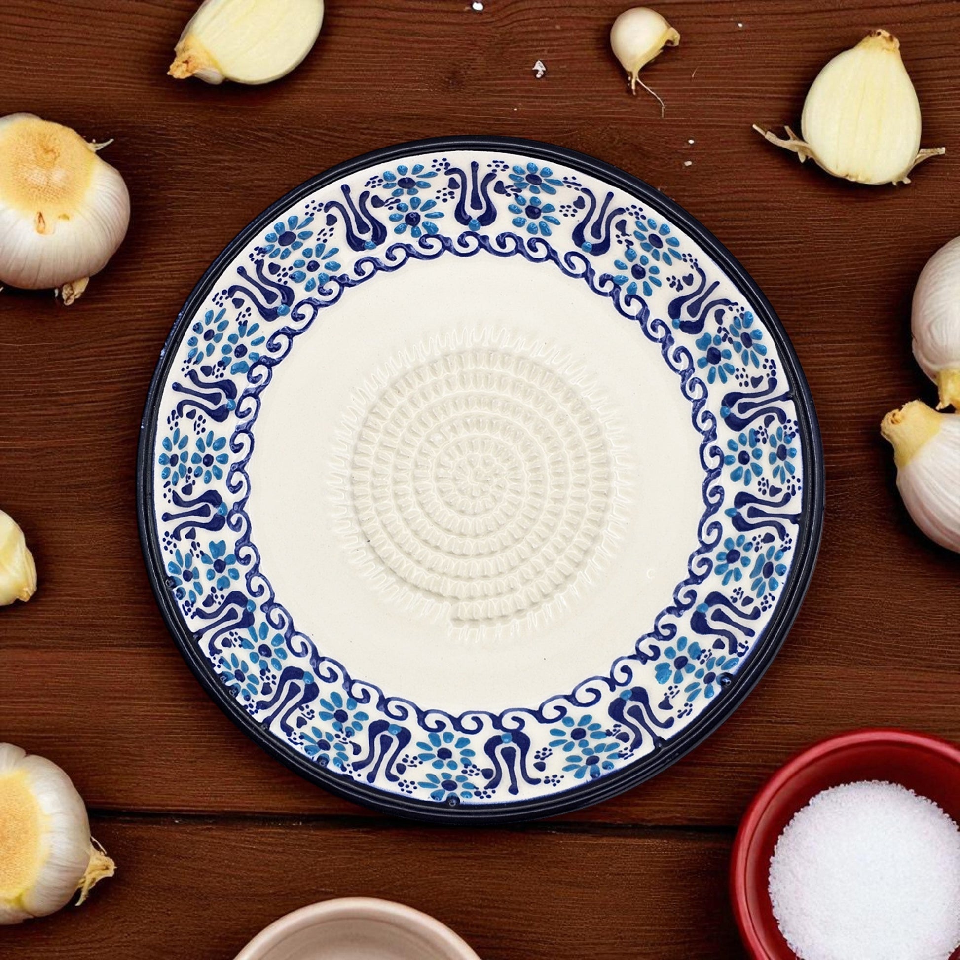 Decorative ceramic garlic grater plate with blue and white pattern on a wooden surface with garlic bulbs.
