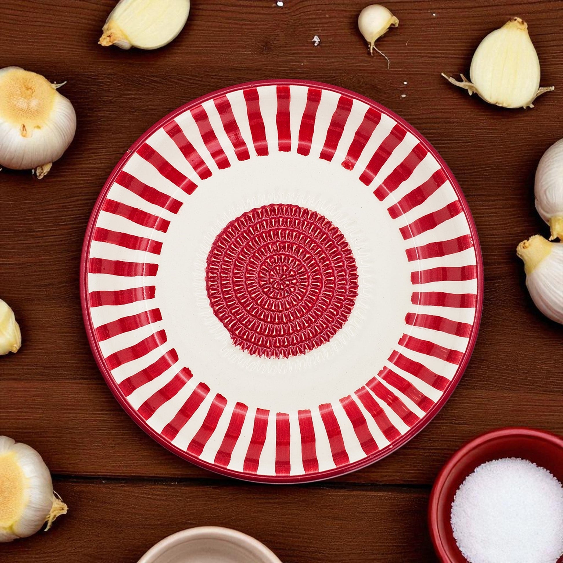 Red and white striped garlic grater bowl on a wooden surface with garlic bulbs and a bowl of salt.