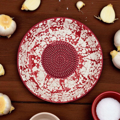 Red and white ceramic garlic grater bowl with decorative center on a wooden surface with garlic bulbs.