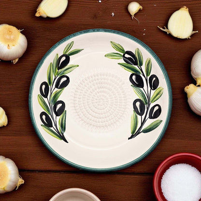 Decorative garlic grater bowl with olive design on a wooden surface with garlic bulbs and a bowl of salt.