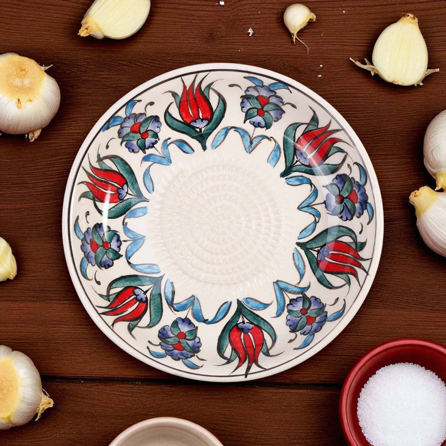 Decorative ceramic garlic grater bowl with floral patterns on a wooden surface with garlic bulbs.