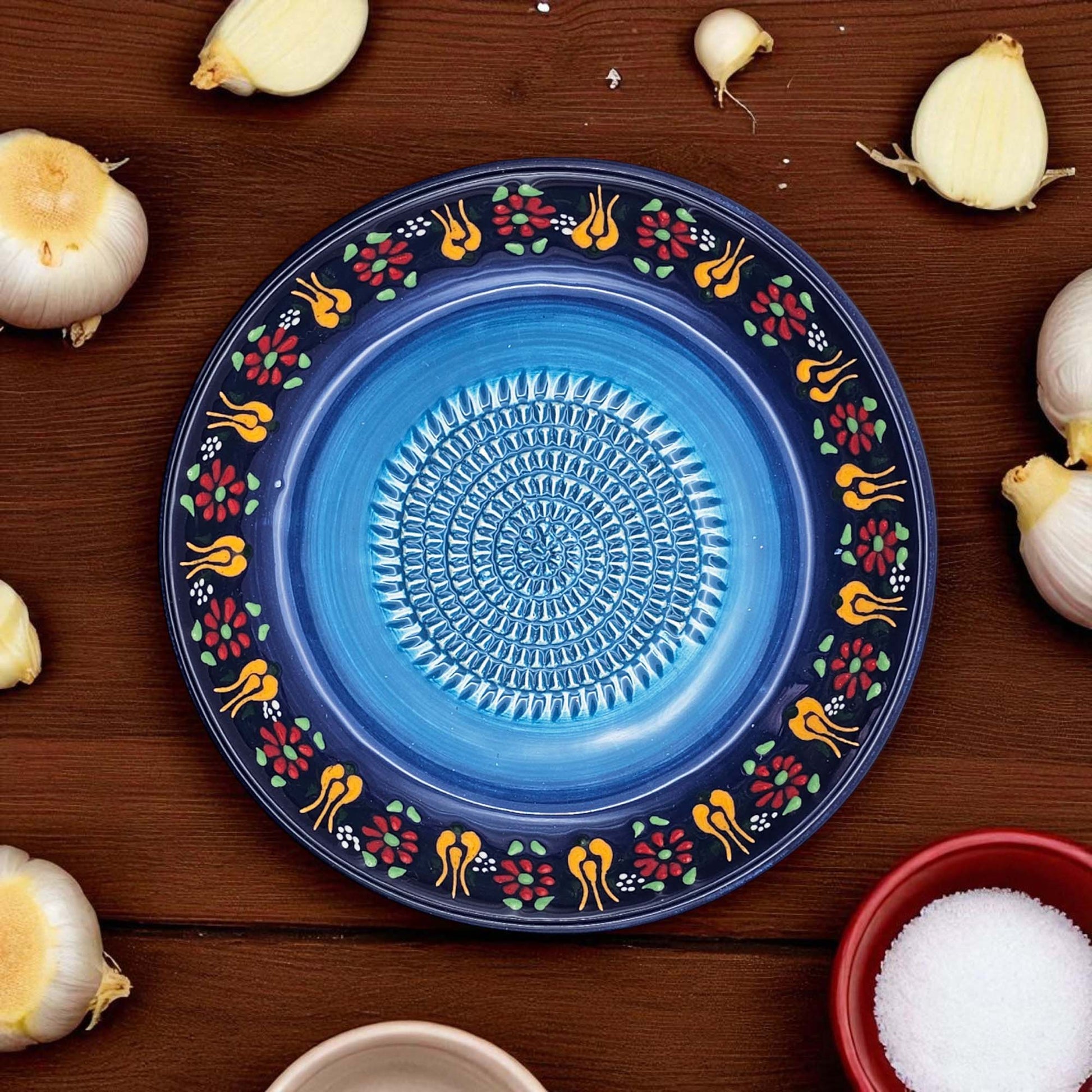 Decorative blue garlic grater bowl with floral patterns on a wooden surface with garlic bulbs and a bowl of salt.