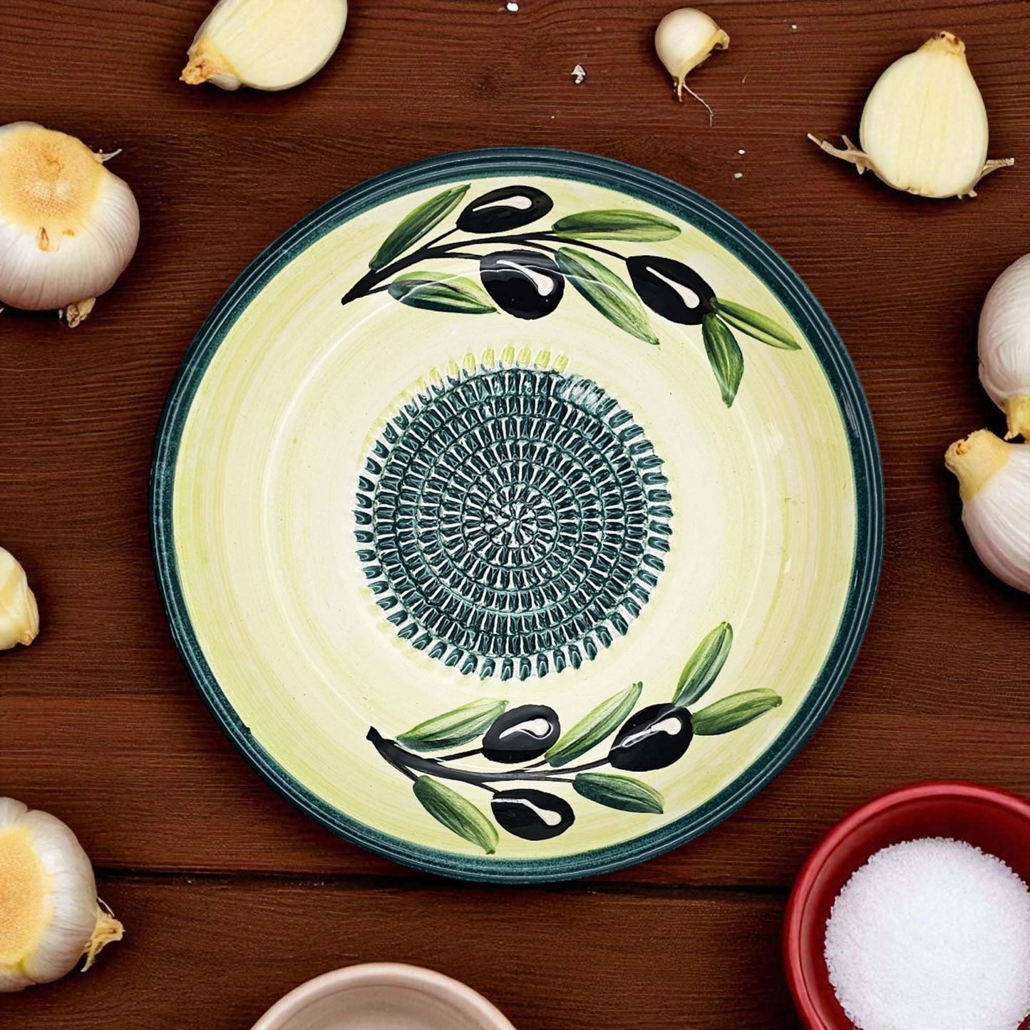 Decorative ceramic garlic grater bowl with olive design on a wooden surface with garlic bulbs.