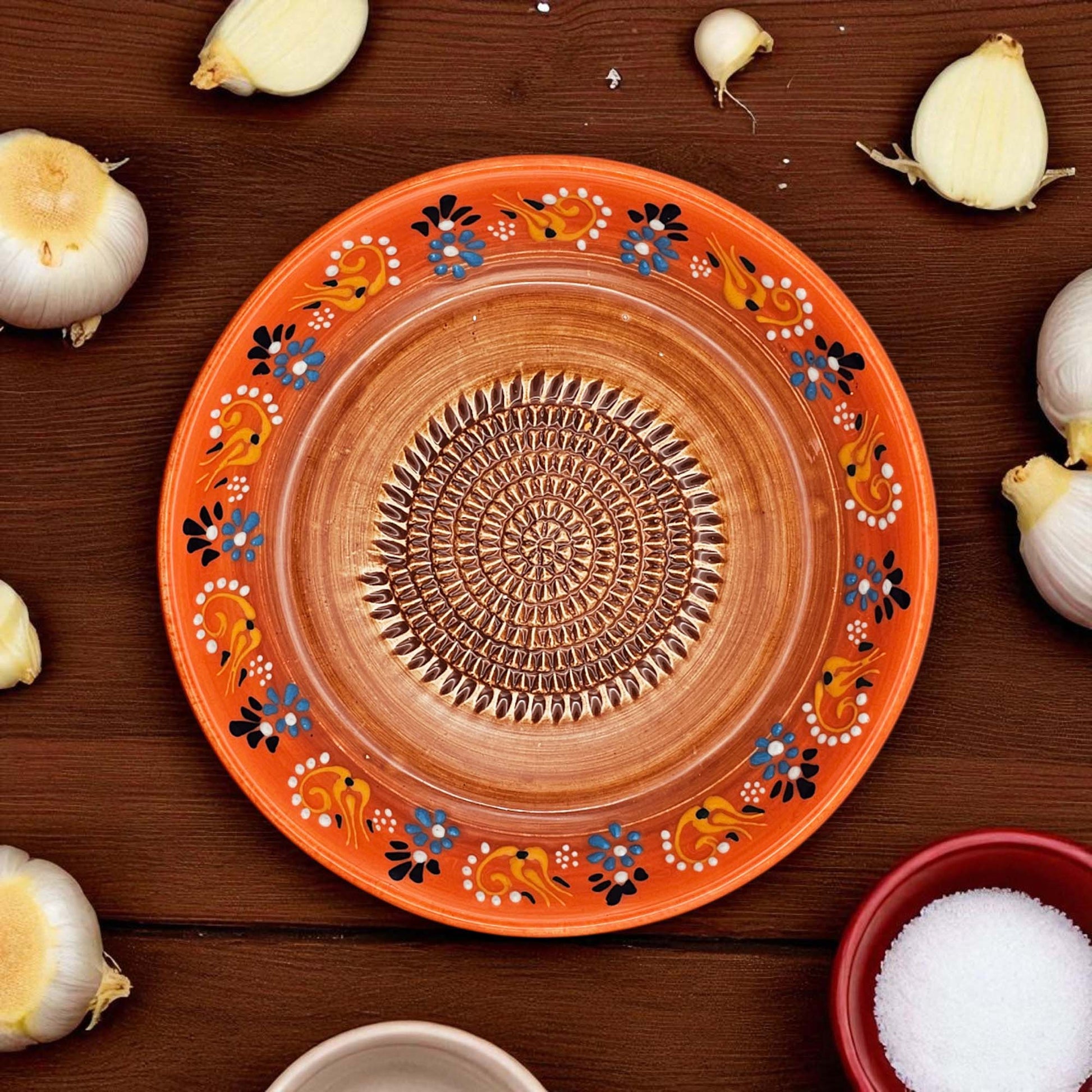 Decorative ceramic garlic grater bowl with floral patterns on a wooden surface with garlic bulbs and a bowl of salt.
