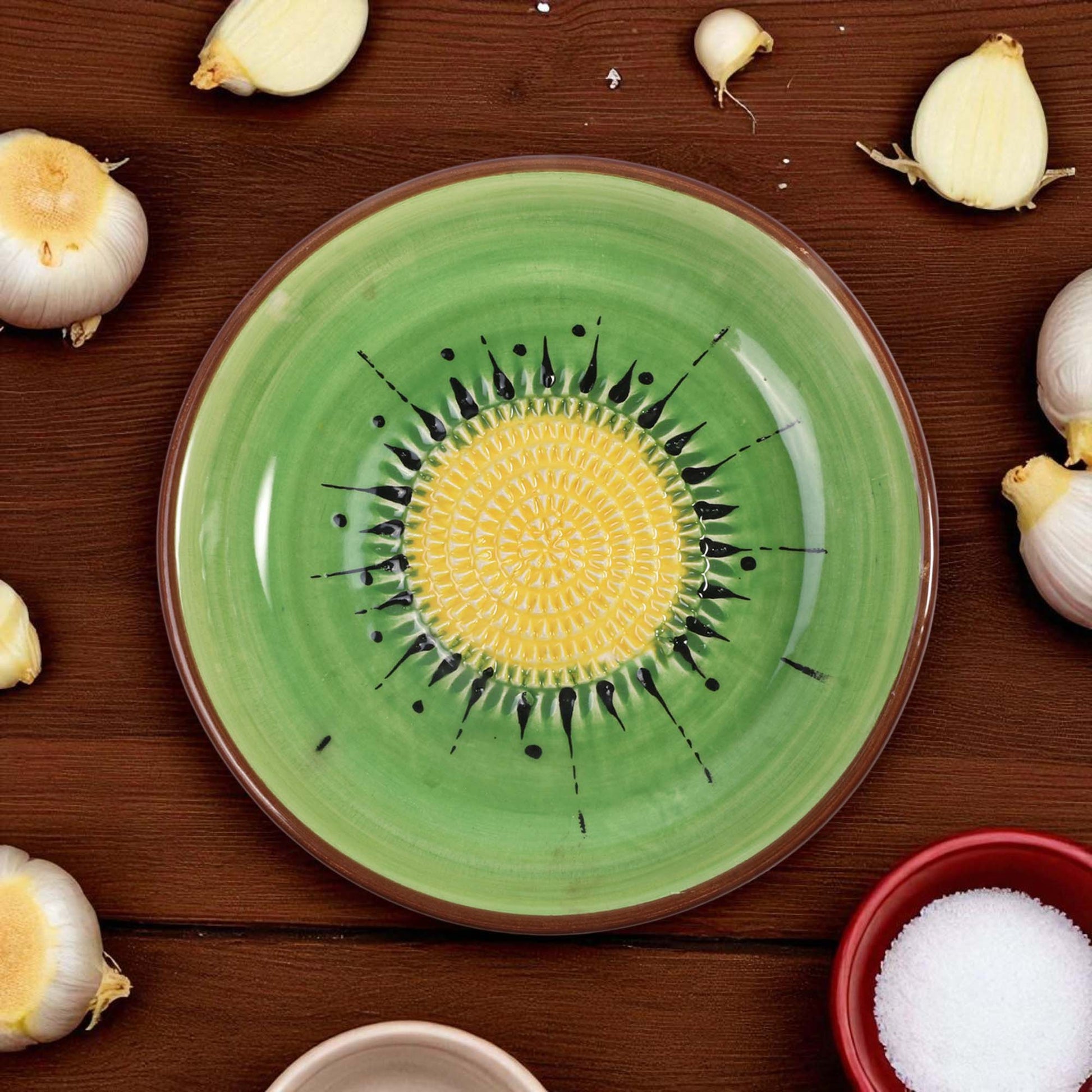 Green ceramic garlic grater bowl with yellow center and black patterns on a wooden surface with garlic bulbs.