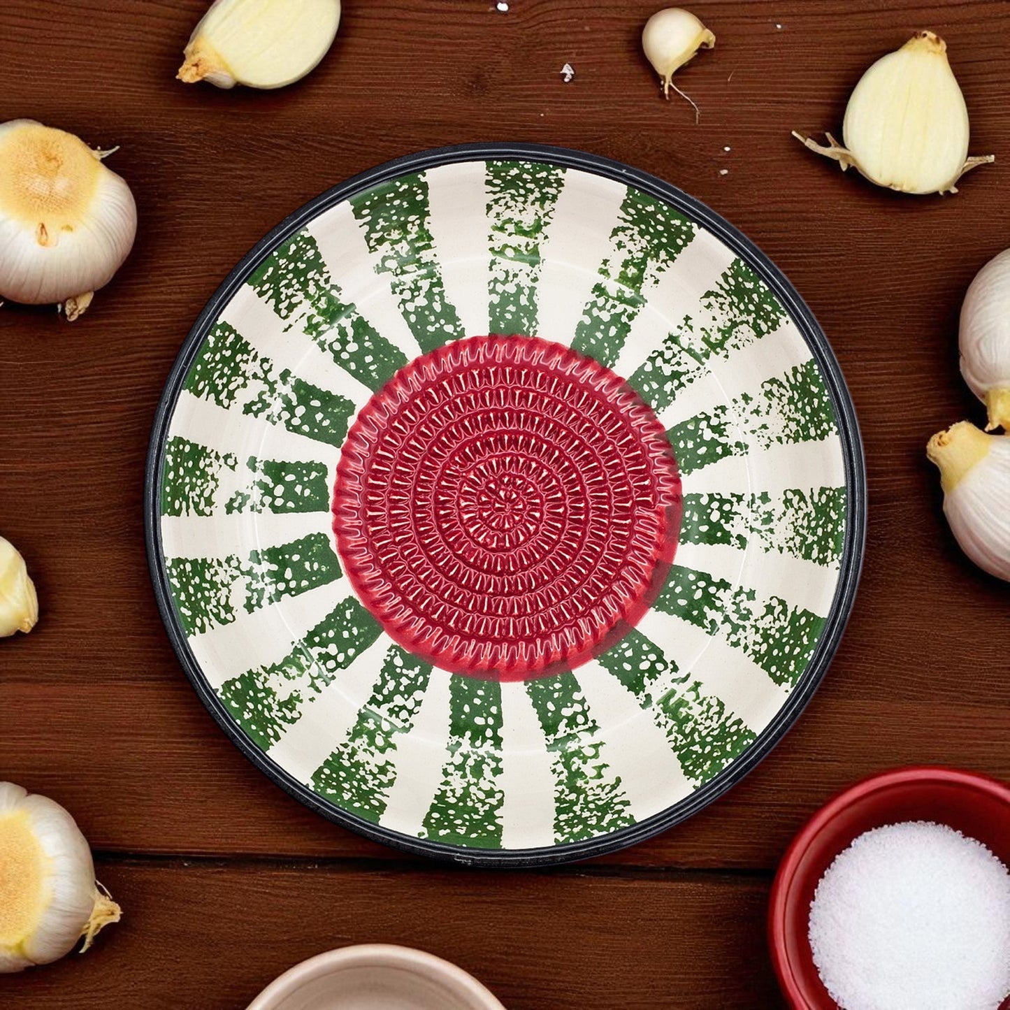 Decorative garlic grater bowl with a red center and green and white pattern on a wooden surface with garlic bulbs.