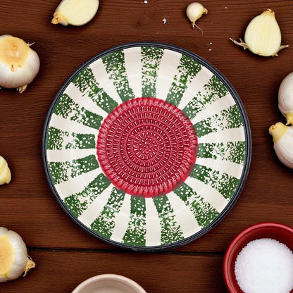 Decorative garlic grater bowl with a red center and green and white pattern on a wooden surface with garlic bulbs.