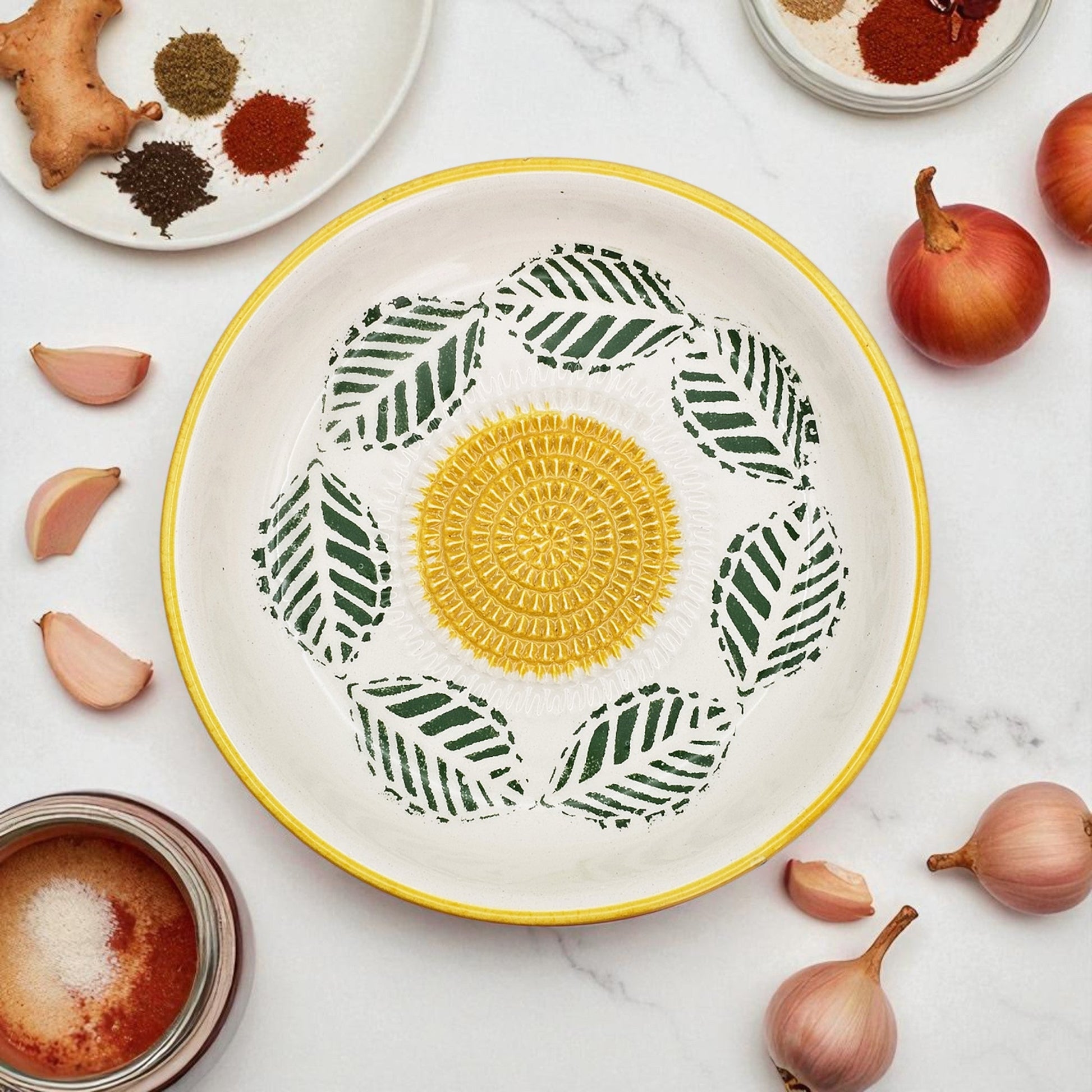 Decorative garlic grater bowl with a yellow center and green leaf patterns on a marble surface with ingredients.