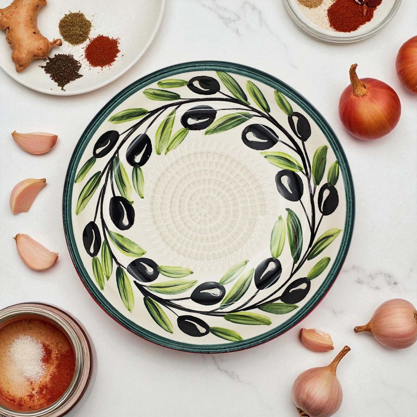 Decorative ceramic garlic grater bowl with olive design on a white surface with ingredients.
