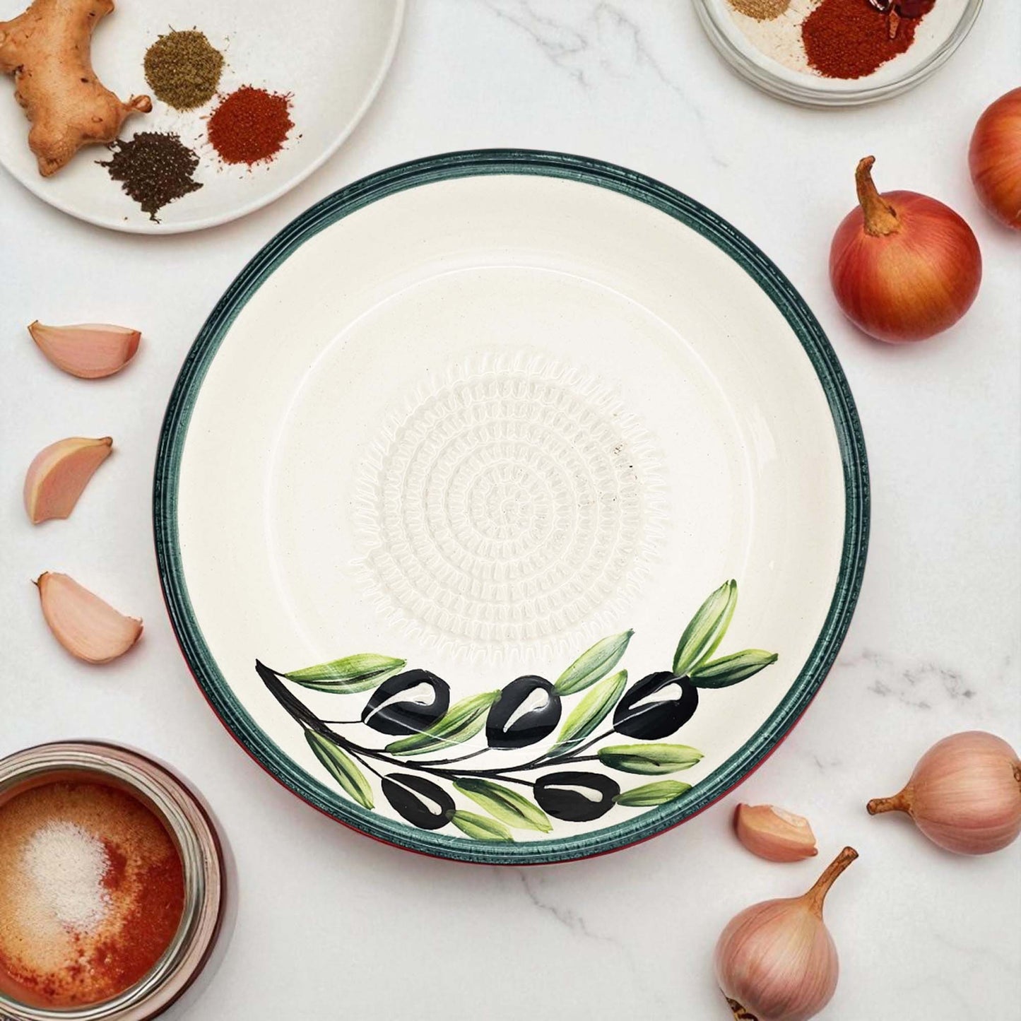 Decorative ceramic garlic grater bowl with olive design on a marble surface with ingredients.