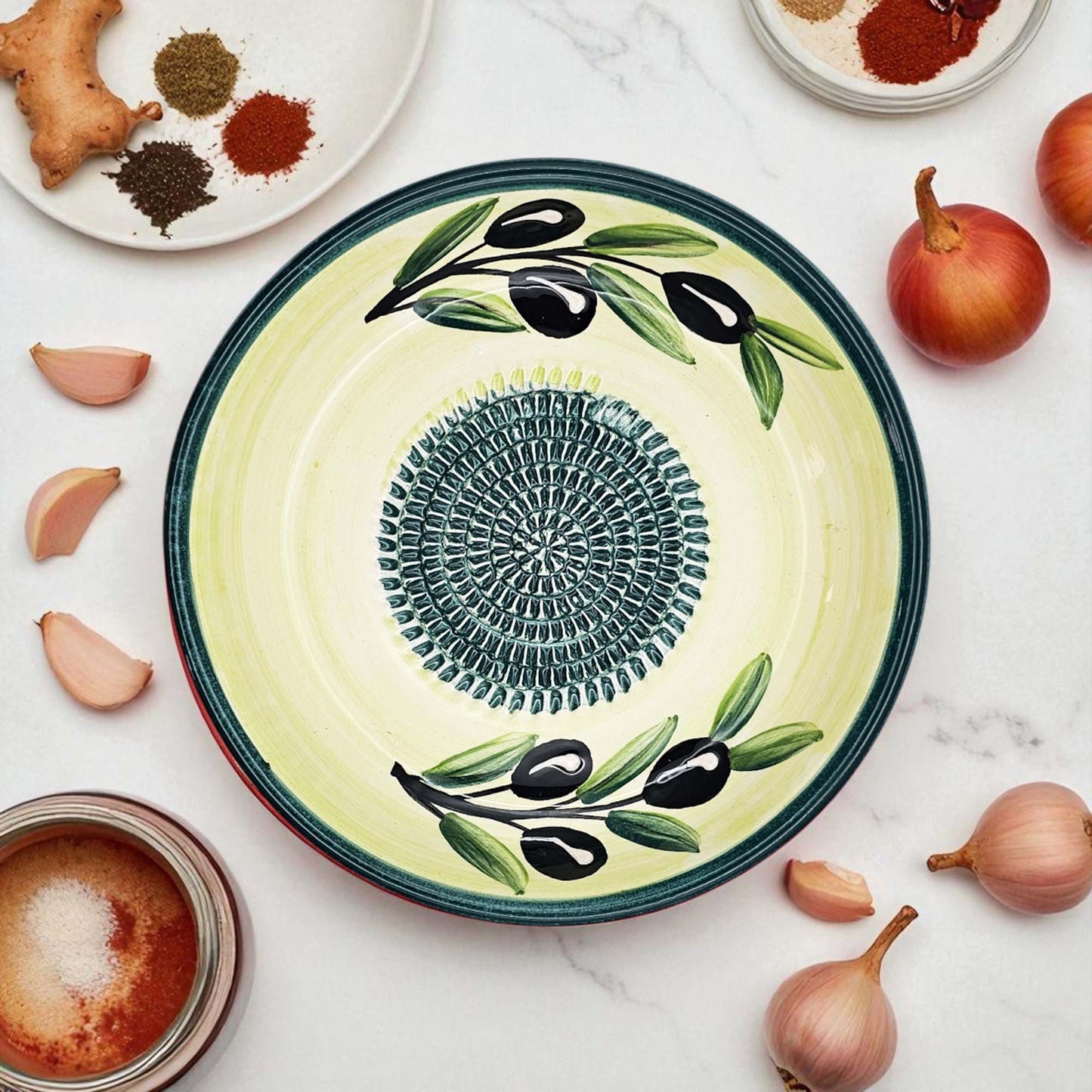 Decorative ceramic garlic grater bowl with olive design on a marble surface with ingredients.