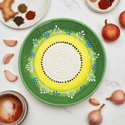 Decorative ceramic garlic grater bowl with green rim and yellow center on a marble surface with ingredients.