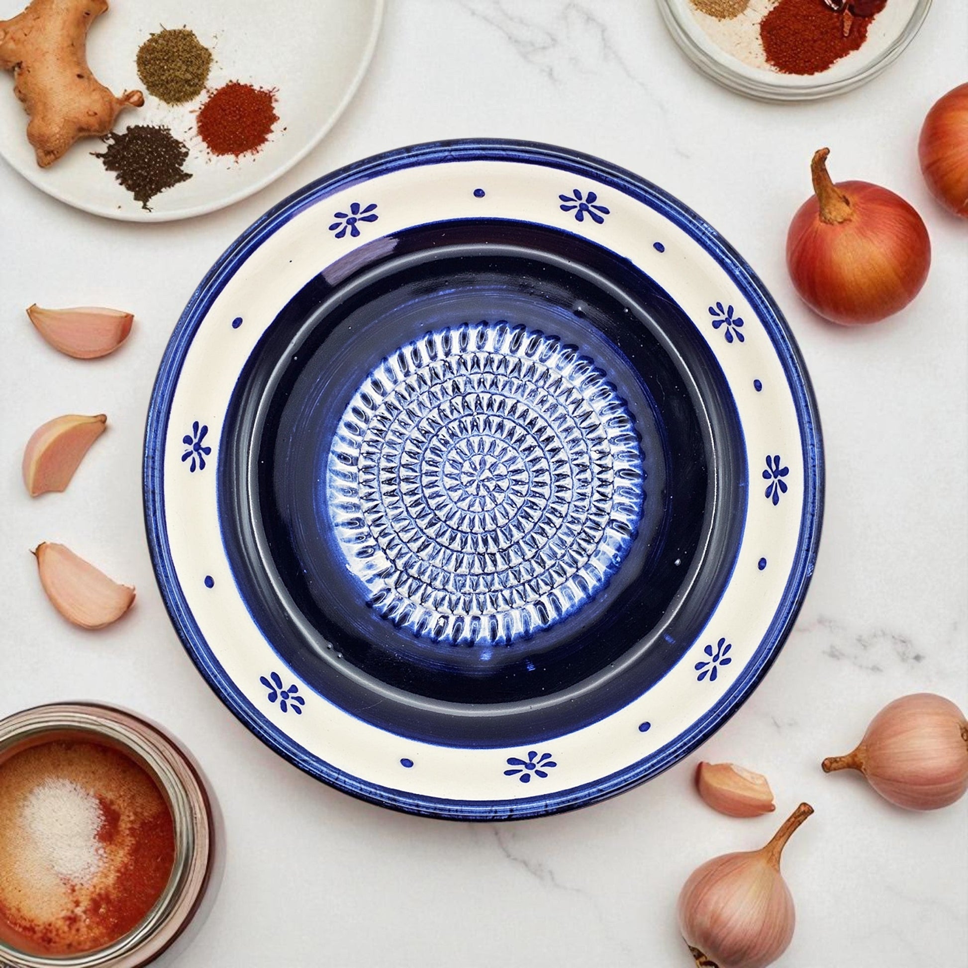 Ceramic garlic grater bowl with blue interior and decorative exterior on a marble surface with ingredients.