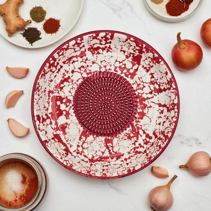 Red and white decorative garlic grater bowl on a marble surface with ingredients around it.