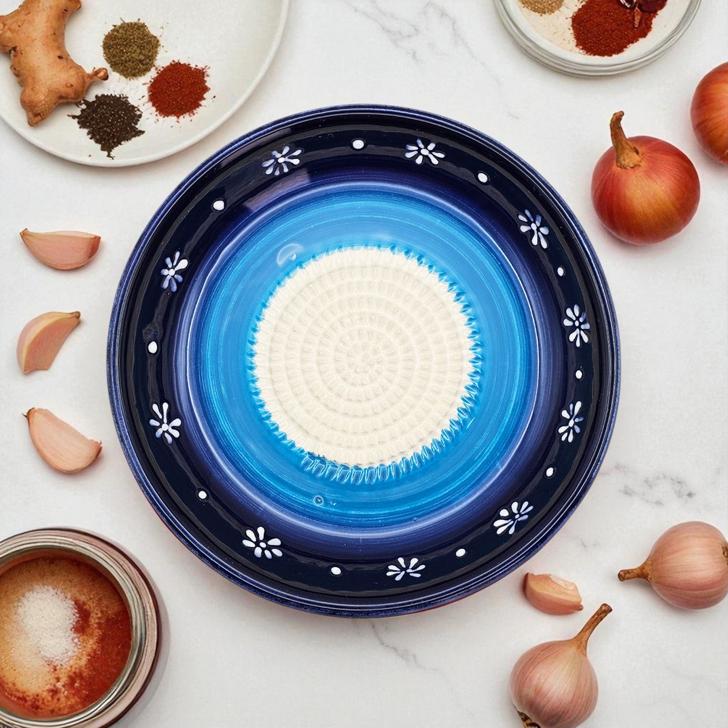 Blue ceramic garlic grater bowl with white center on a marble surface with spices and onions.
