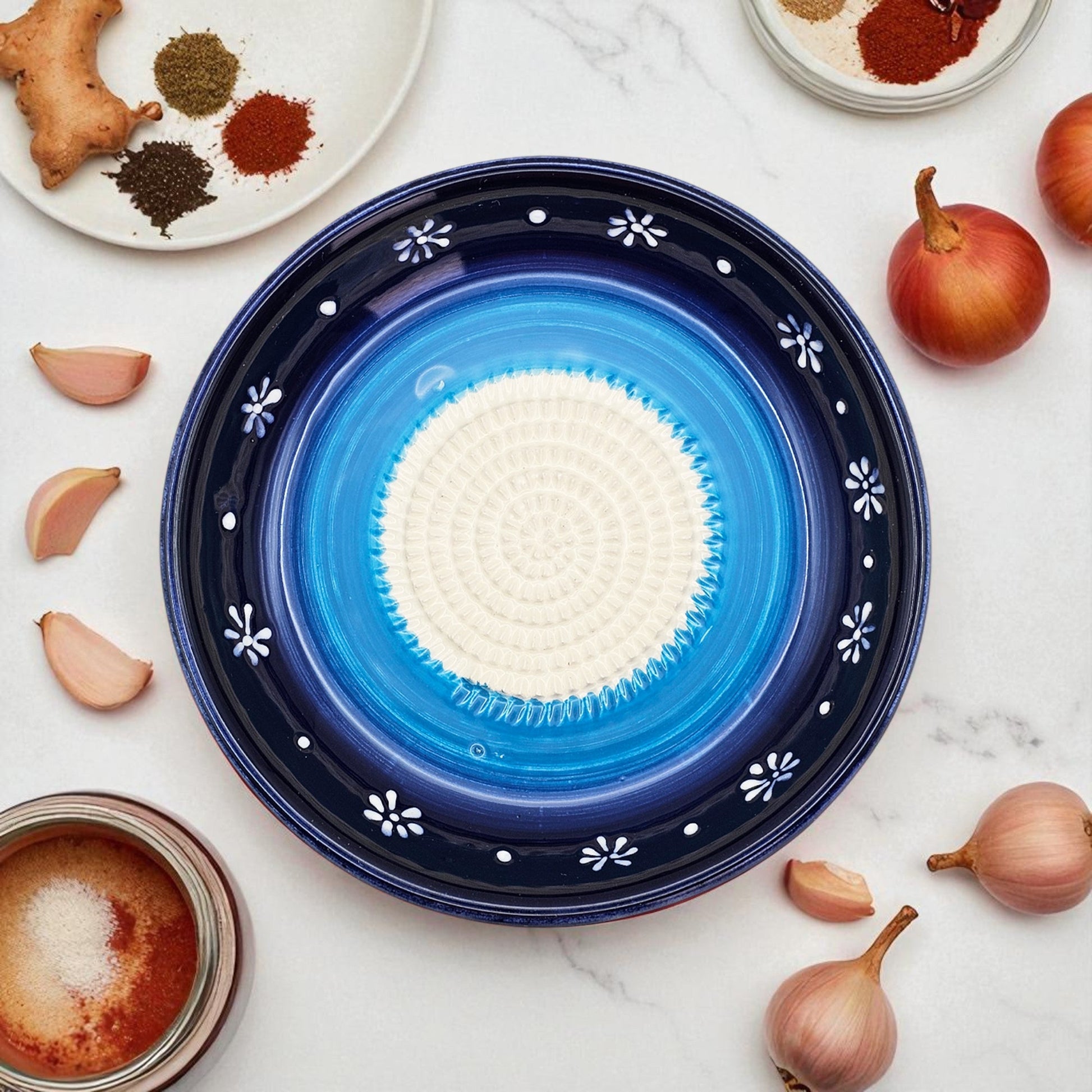 Blue ceramic garlic grater bowl with white center on a marble surface with spices and onions.
