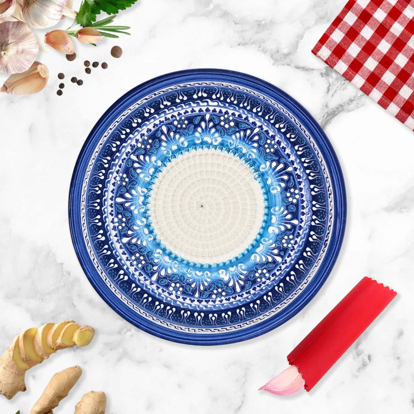 Decorative blue garlic grater bowl on a marble surface with ginger, garlic, and a red checkered cloth.