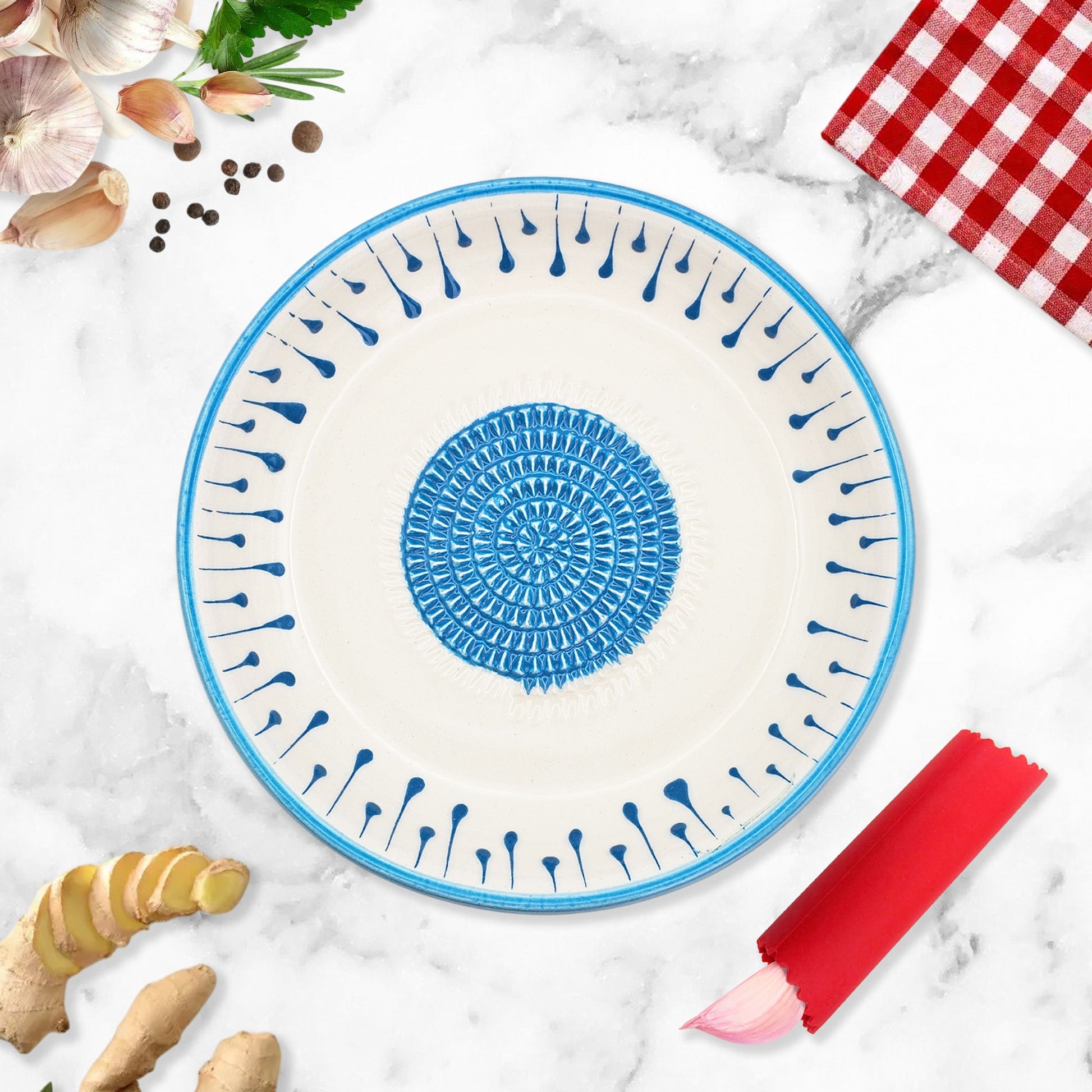 Ceramic garlic grater bowl with blue decorative pattern on a marble surface with ginger and a red checkered cloth.