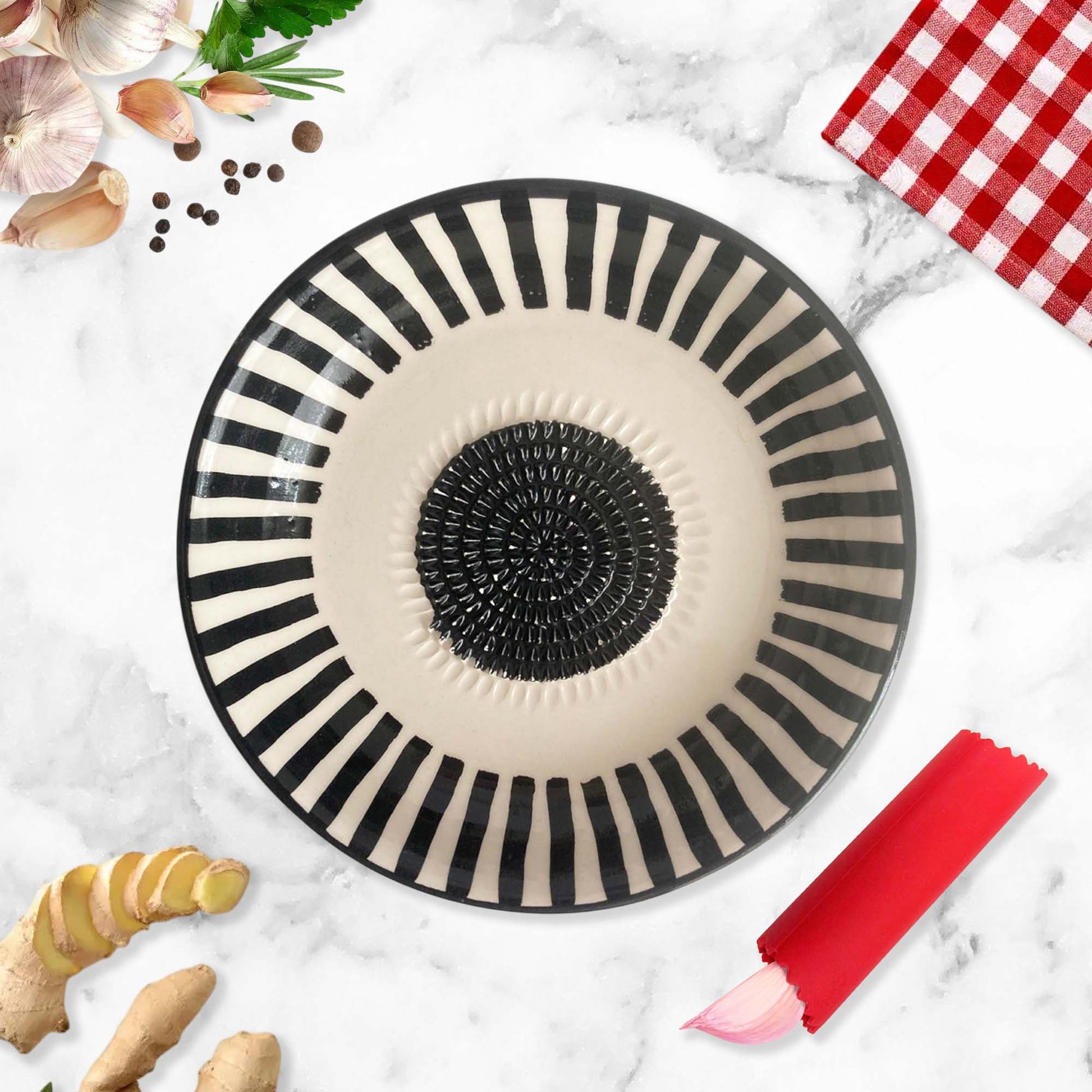 Black and white striped garlic grater bowl on a marble surface with ginger and a red checkered cloth.
